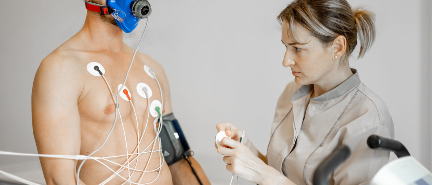 Nurse attaches electrodes to a man for a cardio endurance test during physical exercise on bike simulator, examining heart and vascular system. Man in breath mask on face