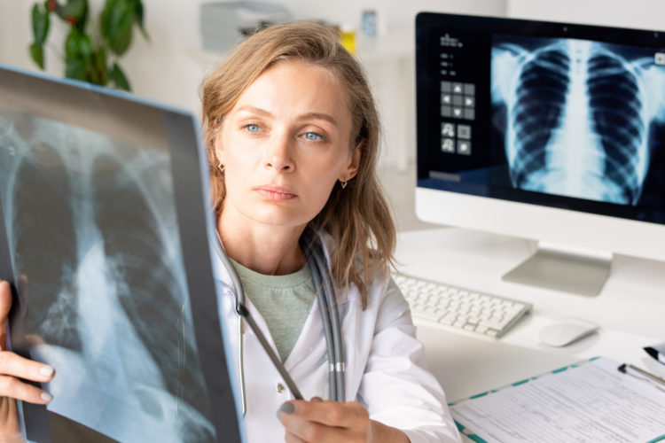 Young serious female radiologist in whitecoat pointing at x-ray image of lungs